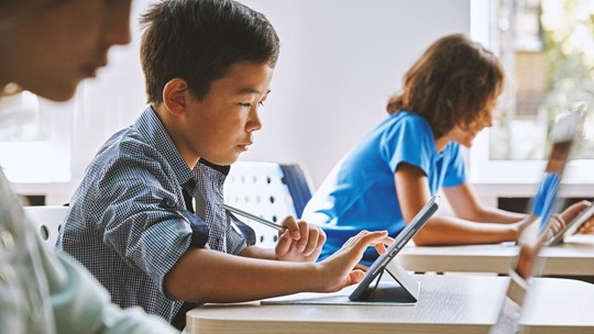 Focused Asian school boy using digital tablet at class in classroom. Attentive junior school student learning online virtual education digital program app tech during stem computer science lesson.